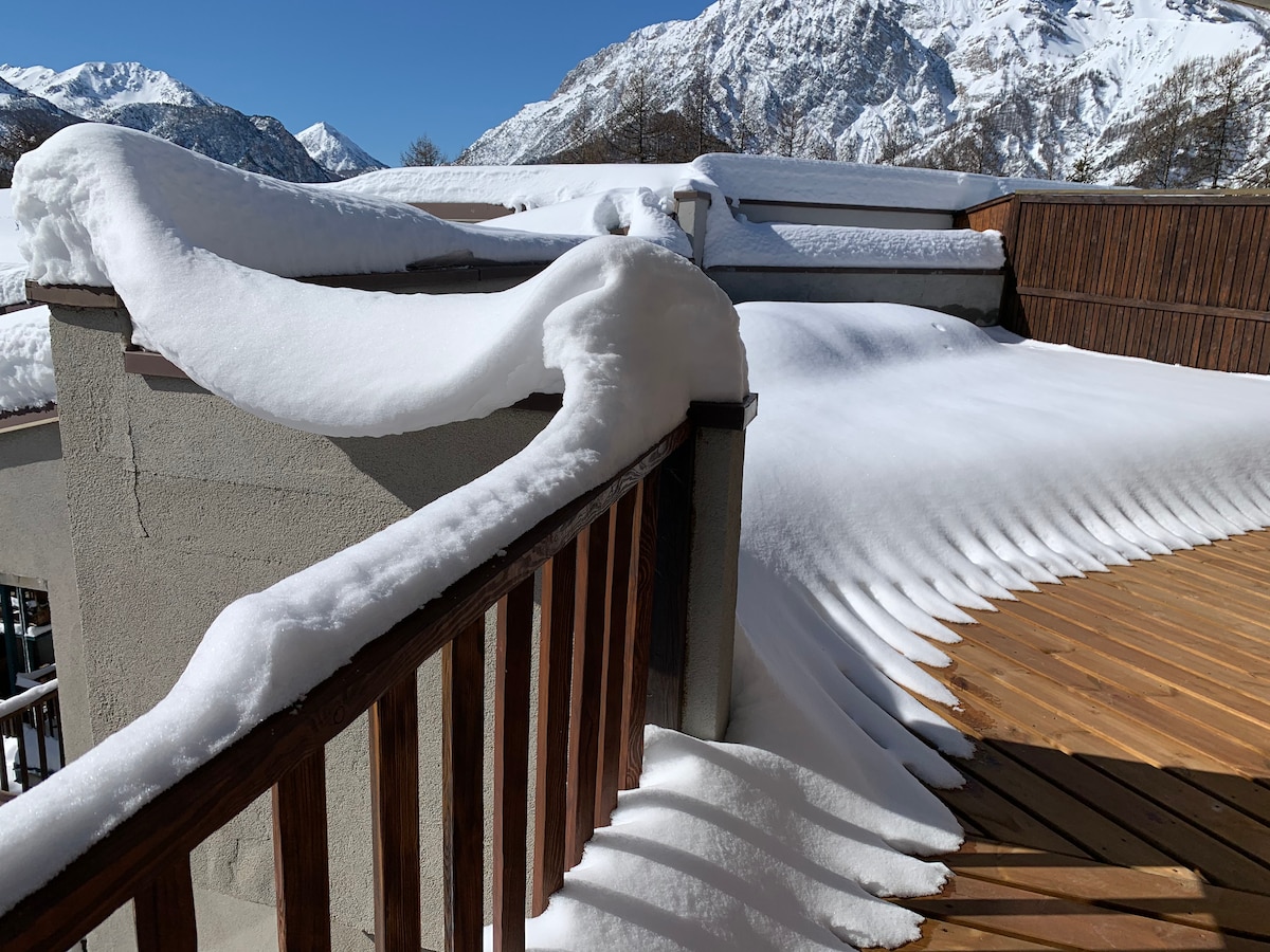 A large terrace with wooden flooring is covered in a soft layer of fresh snow. Snow gently cascades over the edge of a low wall, while a clear blue sky and mountain peaks create a serene backdrop.