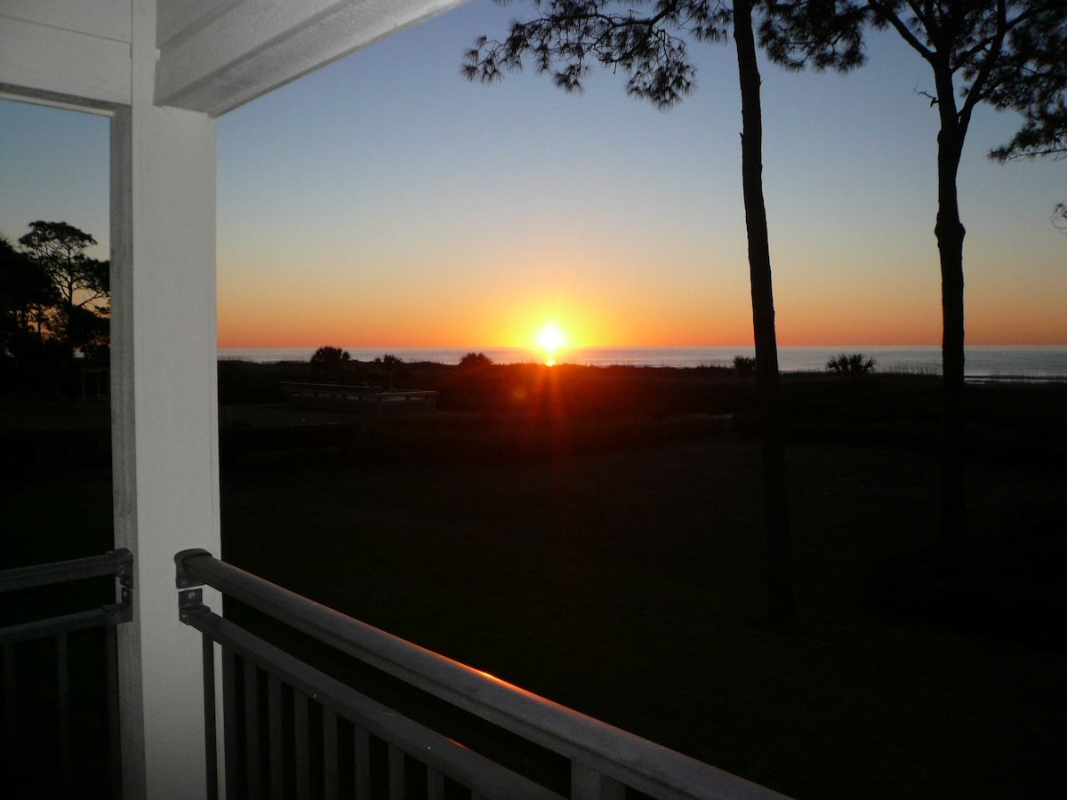A serene view of a sunset over the ocean is framed by a railing, with silhouettes of trees in the foreground. The horizon displays vibrant orange and yellow hues against a deepening blue sky, providing a tranquil backdrop to the surrounding landscape.