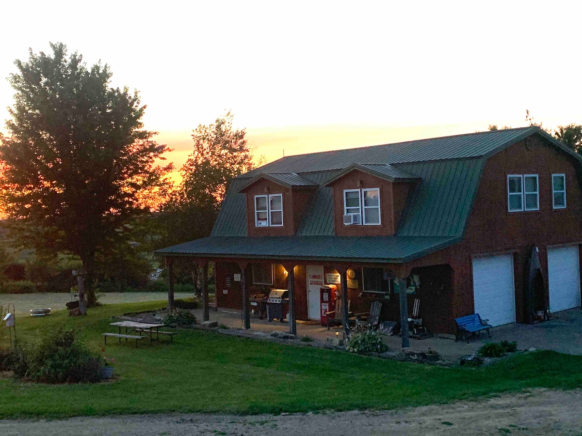 The exterior of the house is shown at sunset, featuring a rustic design with a green metal roof. A porch is visible, furnished with seating and a grill, set against a backdrop of expansive grass and trees, creating a calm rural atmosphere.