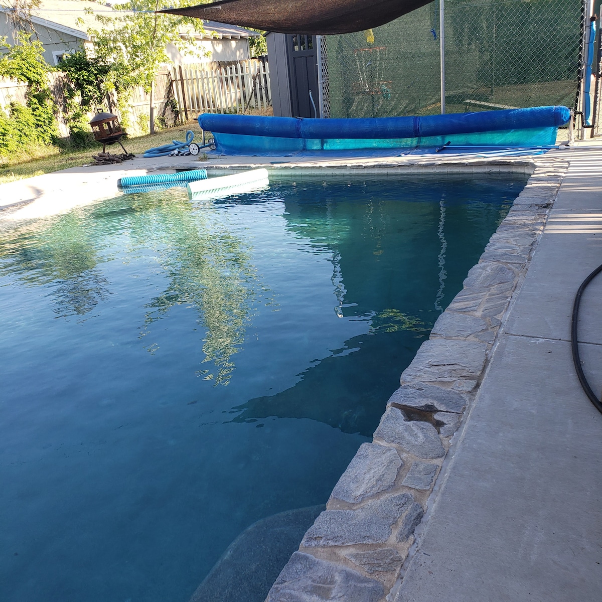 A refreshing swimming pool is surrounded by a stone deck, with a blue cover partially visible. Lush greenery is present in the background, providing a natural setting. The pool's clear water reflects the sunlight, creating a tranquil atmosphere.