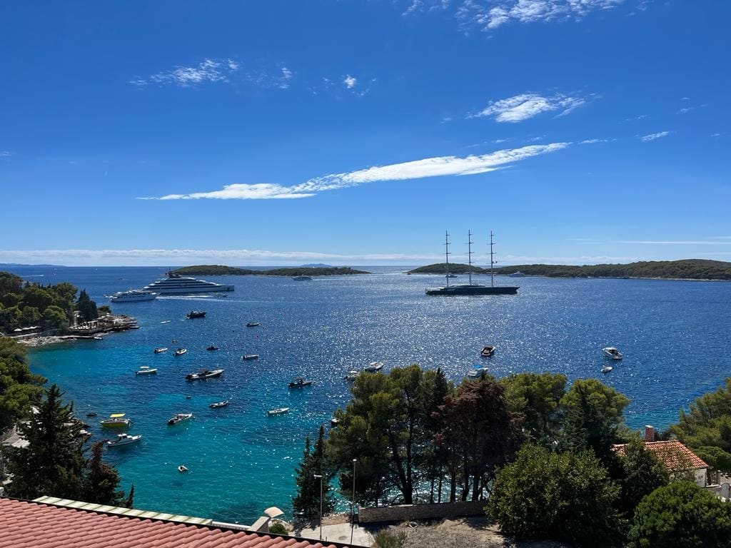 A scenic view of the sea and islands is presented, featuring clear blue waters and a variety of boats. The coastline is lined with greenery, while the sky is bright with a few wispy clouds, creating a serene and inviting atmosphere.