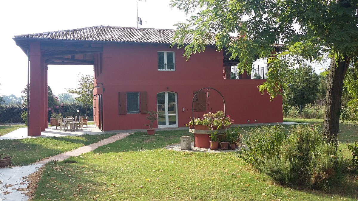 A red two-story building is surrounded by greenery, with large windows and a covered outdoor area. A pathway lined with potted plants leads to a patio furnished with a table and chairs, enhancing the inviting outdoor space.