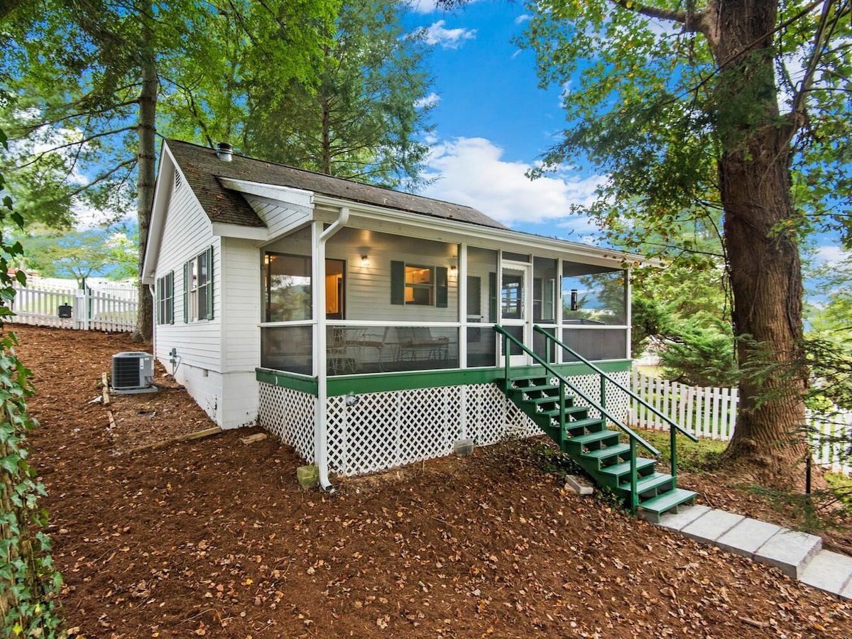 A charming cottage exterior features a screened porch with multiple chairs and a table, surrounded by greenery. Steps lead to the entrance, and a white picket fence outlines the property, providing a cozy and inviting atmosphere in a natural setting.