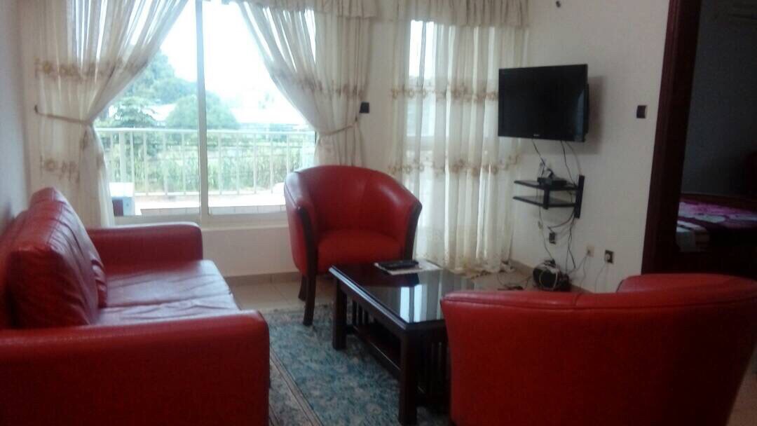 A living room features two bright red armchairs and a matching sofa arranged around a dark wooden coffee table. Natural light filters through sheer curtains hung over a large window, offering views of the outside. A small wall-mounted TV is positioned for entertainment.