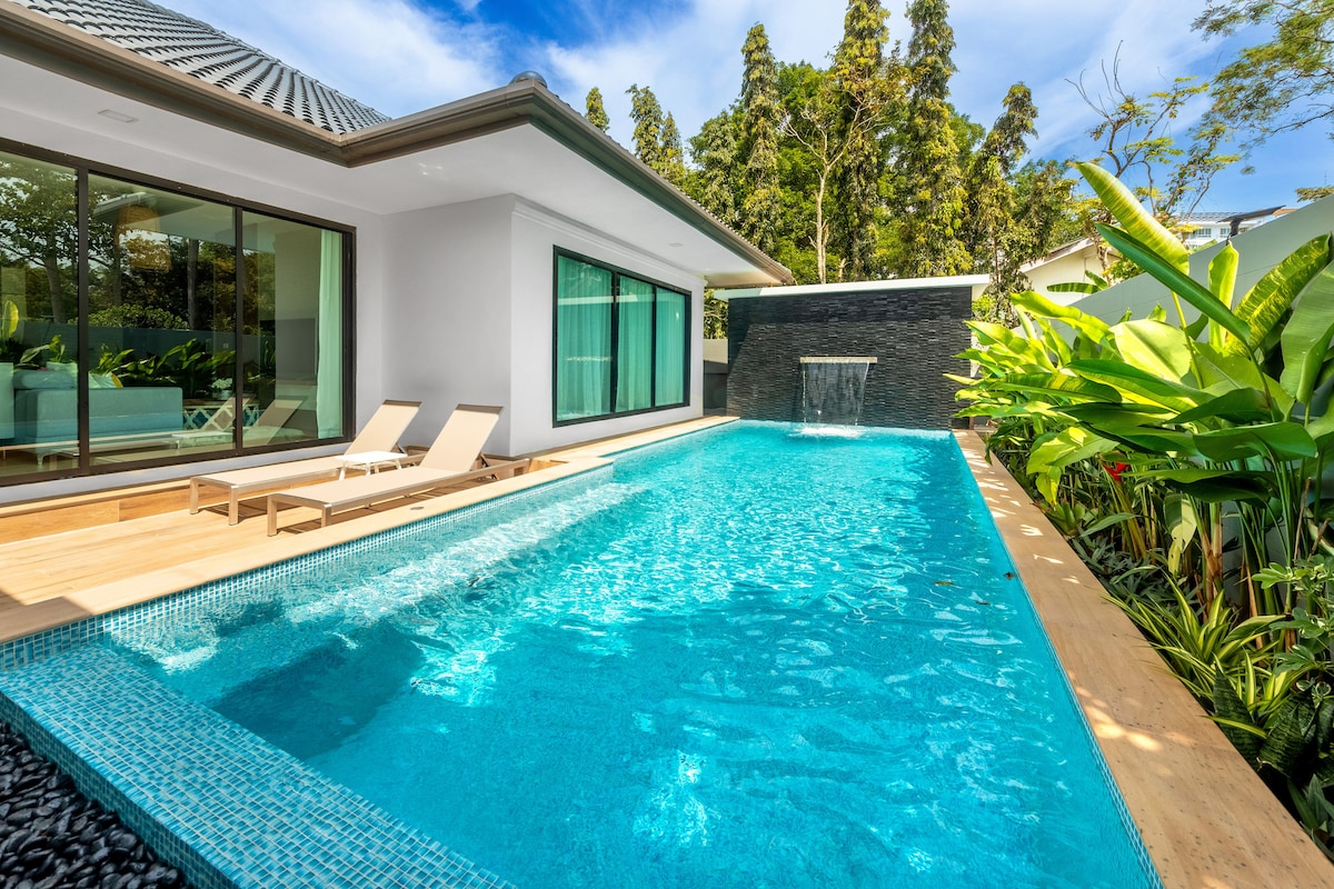 A private swimming pool is surrounded by lush greenery and wooden decking. Sun loungers are positioned beside the water, inviting relaxation. The villa's modern exterior features large glass doors that offer a seamless view of the pool area, enhancing an airy atmosphere.