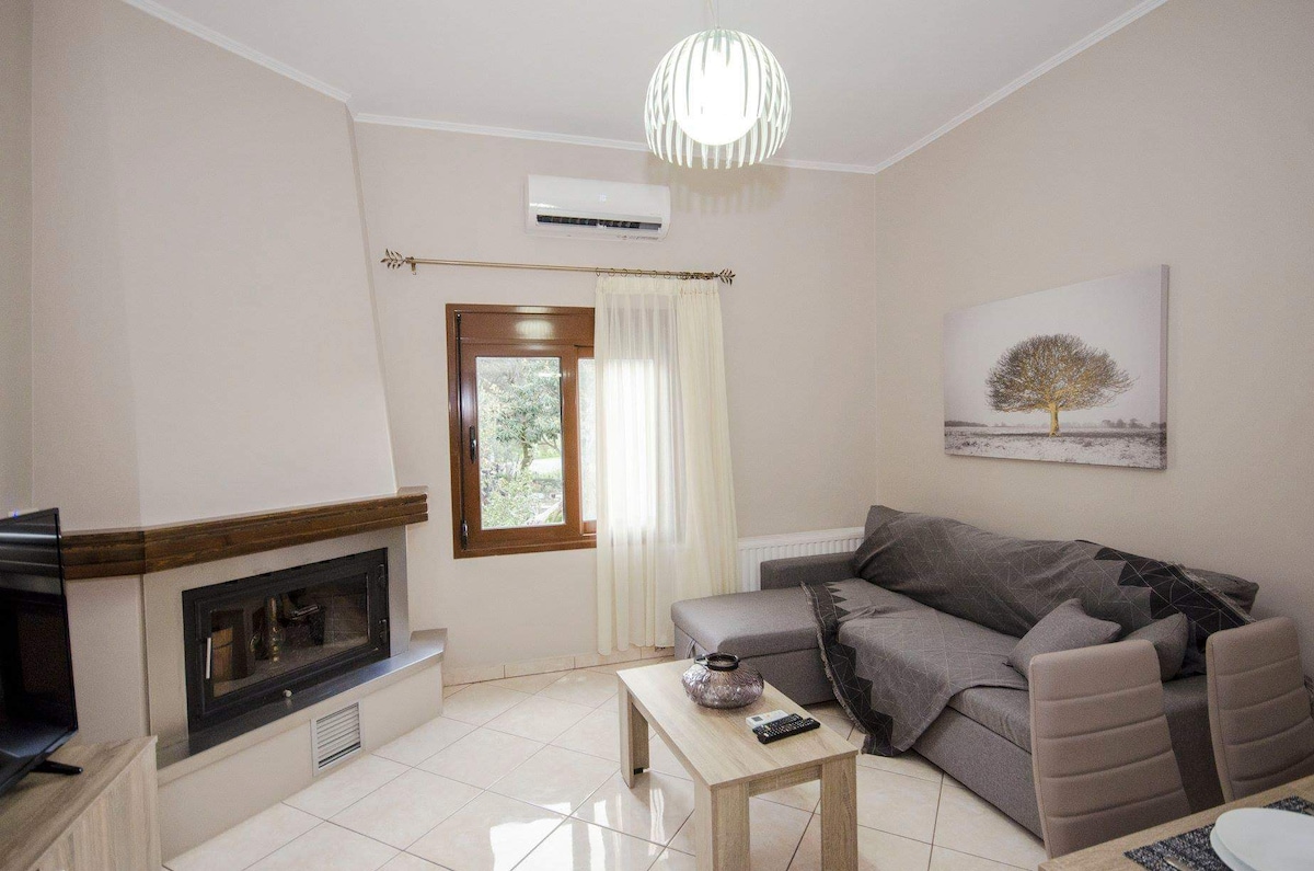 A living area features a comfortable sofa with a cozy blanket draped over it, situated near a fireplace. A wooden table sits in front of the sofa. Natural light enters through a window adorned with light curtains, enhancing the serene ambiance of the room.
