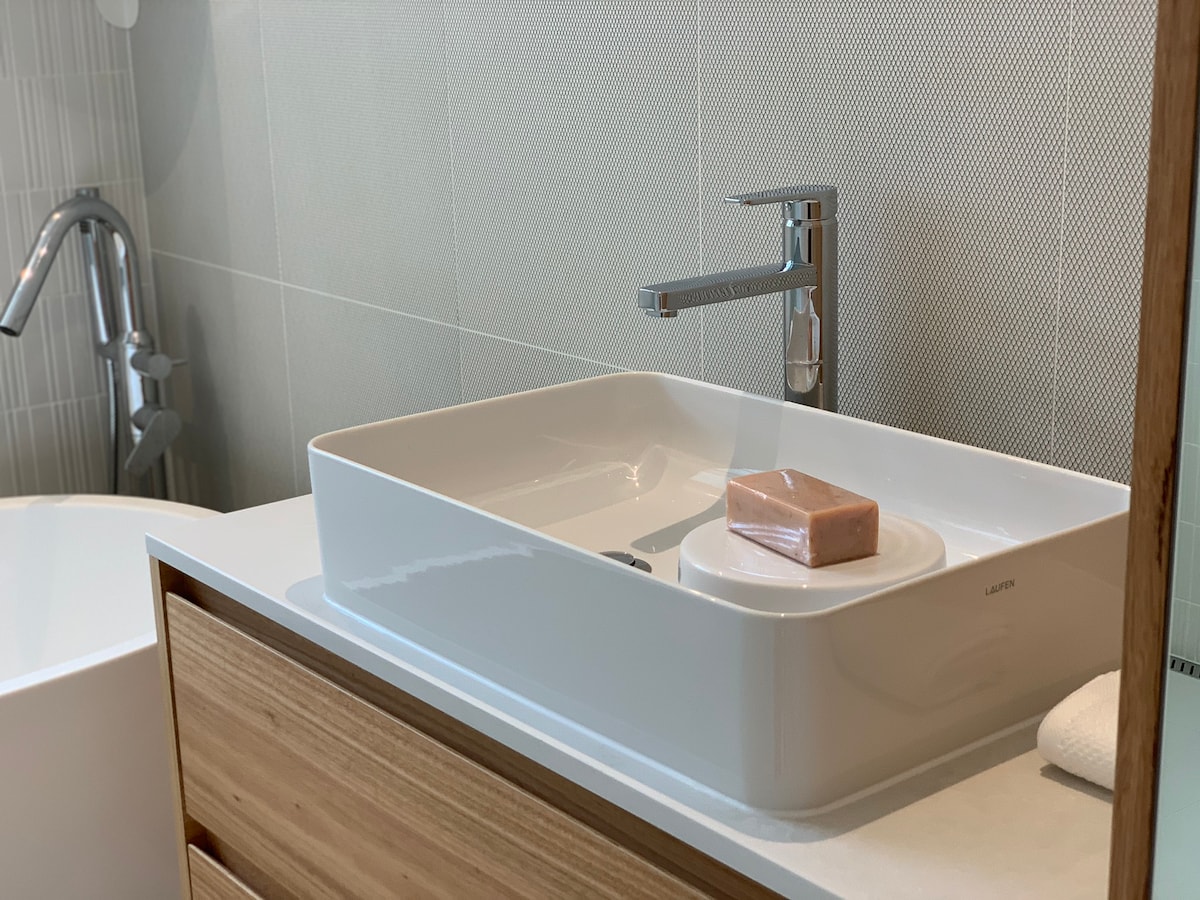 A minimalist bathroom sink setup is displayed, featuring a rectangular basin atop a wooden vanity. A bar of soap rests on a circular dish. The faucet is sleek, and the wall is adorned with textured tiles that complement the overall clean aesthetic.