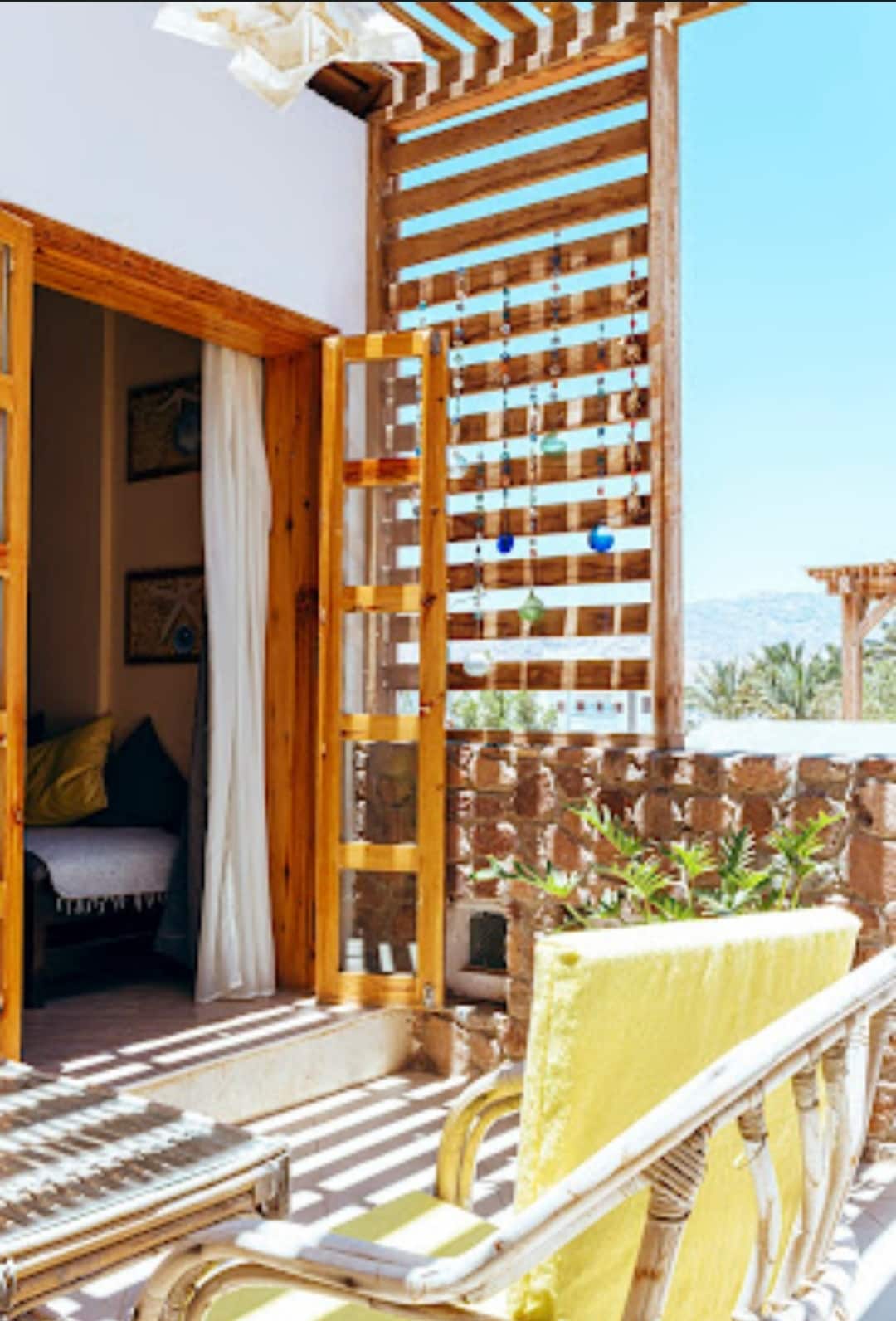 A bright terrace area features wooden slatted walls and bright sunshine filtering through. Two cozy chairs are positioned nearby, and ornamental hanging decorations provide a touch of color. Lush greenery is visible in the background, enhancing the inviting outdoor space.