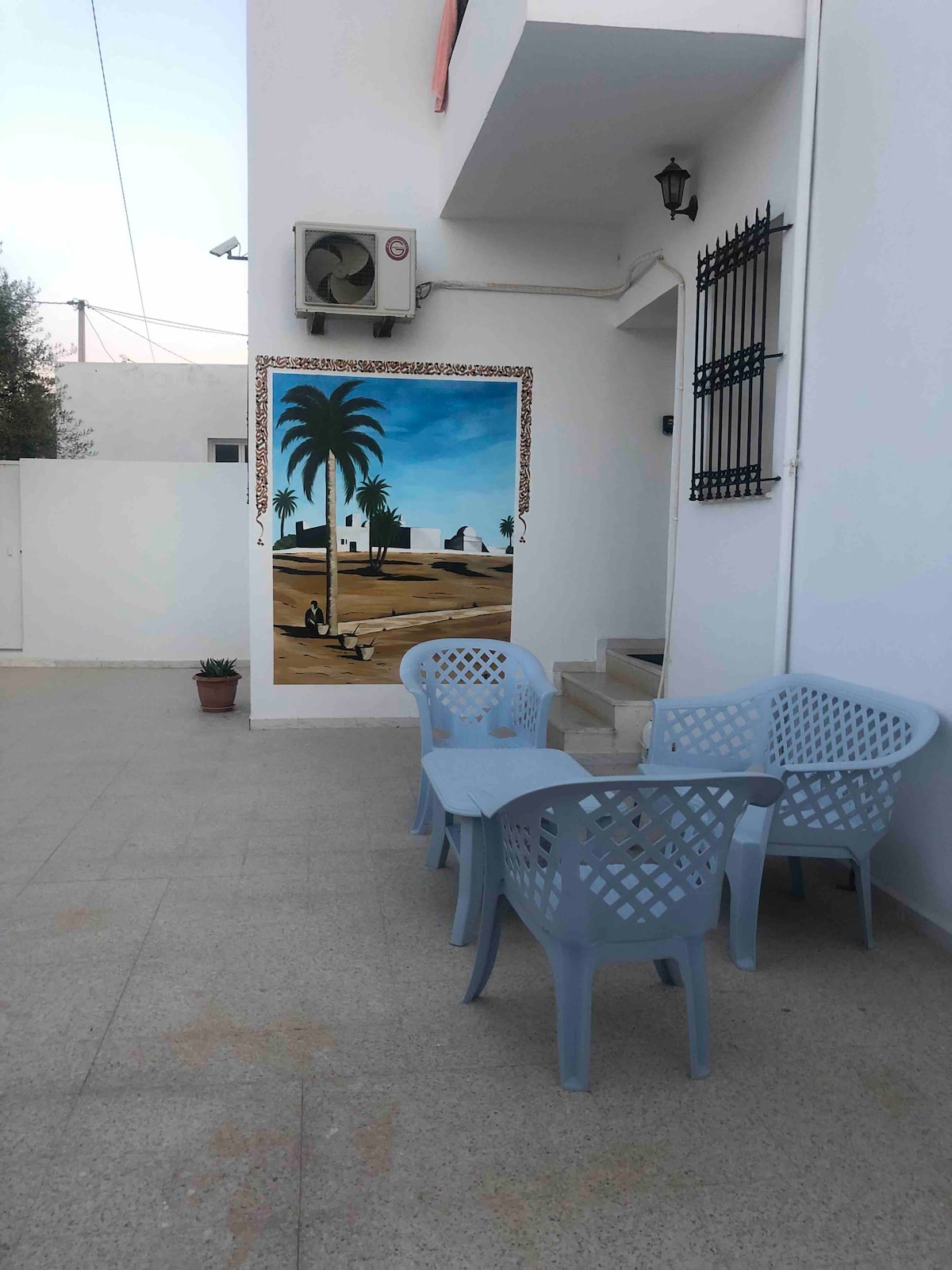 An outdoor seating area is showcased with light blue chairs and a small table, set against a vibrant mural depicting palm trees and a scenic landscape. The paved floor surrounding the area is well-maintained, with a potted plant adding a touch of greenery.
