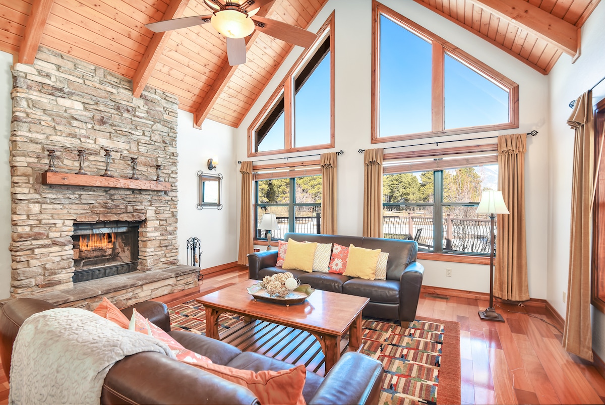 The spacious living area is adorned with high, vaulted wooden ceilings and large windows, allowing natural light to fill the space. A stone fireplace is featured prominently, surrounded by comfortable seating and a wooden coffee table, creating a cozy environment for relaxation.
