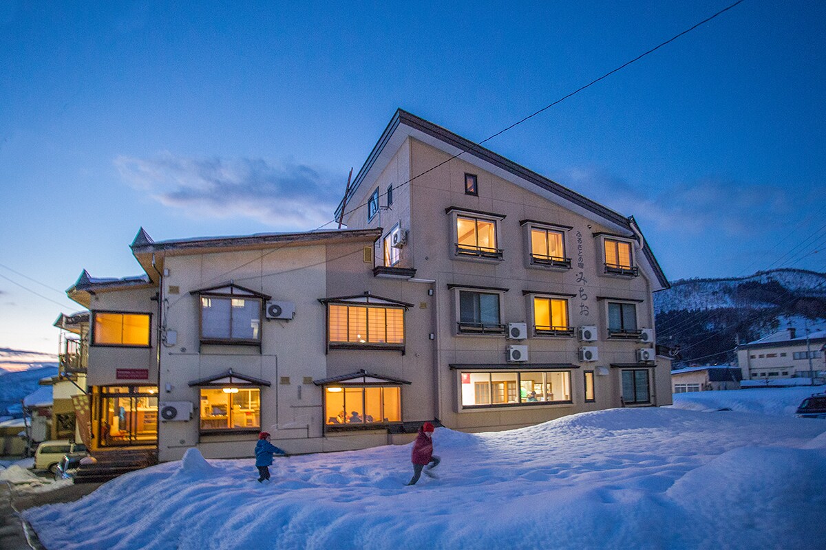 A multi-story building is seen at dusk, with warm light glowing from its windows. Snow blankets the ground, and children can be seen playing in the foreground. The surrounding mountains provide a scenic backdrop against a twilight sky.
