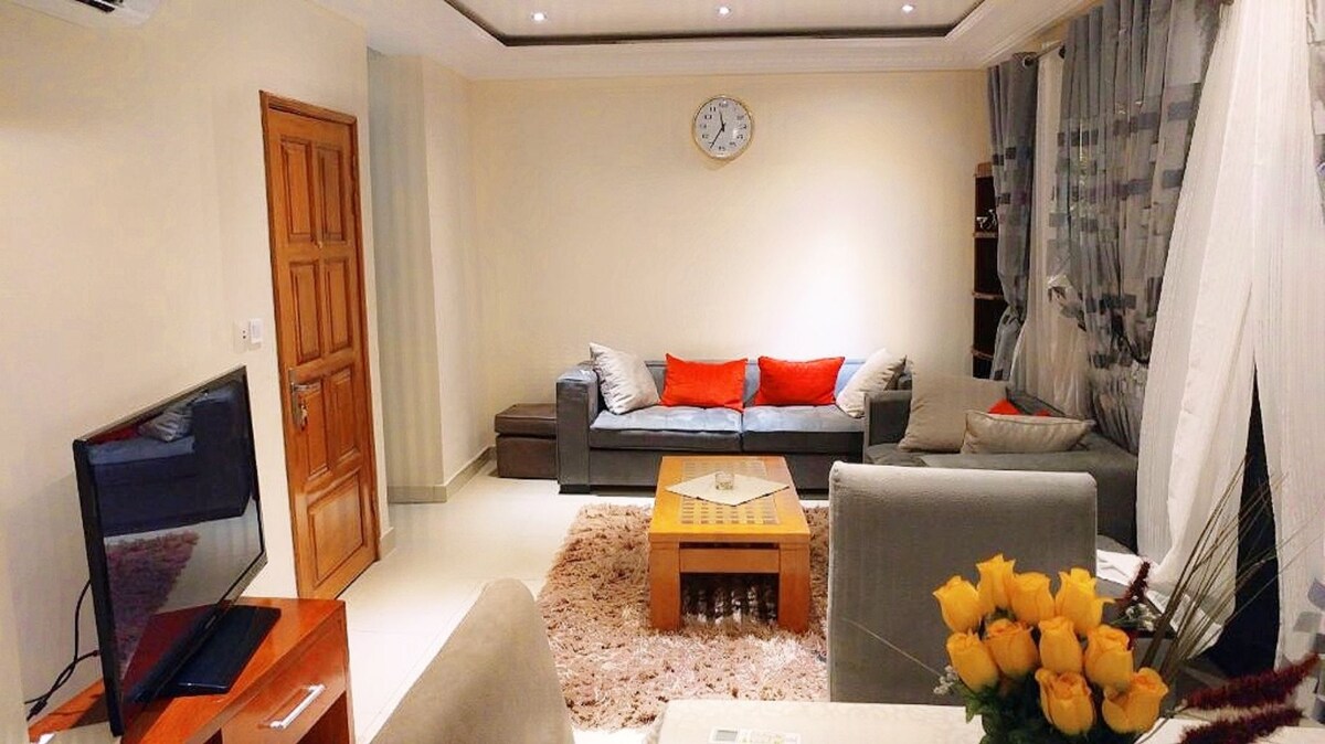 A cozy living room features a gray sectional sofa with contrasting red cushions, paired with a wooden coffee table. Natural light filters through sheer curtains, illuminating a wall-mounted television and decorative accents. A vase of yellow flowers sits on a side table, adding a cheerful touch.