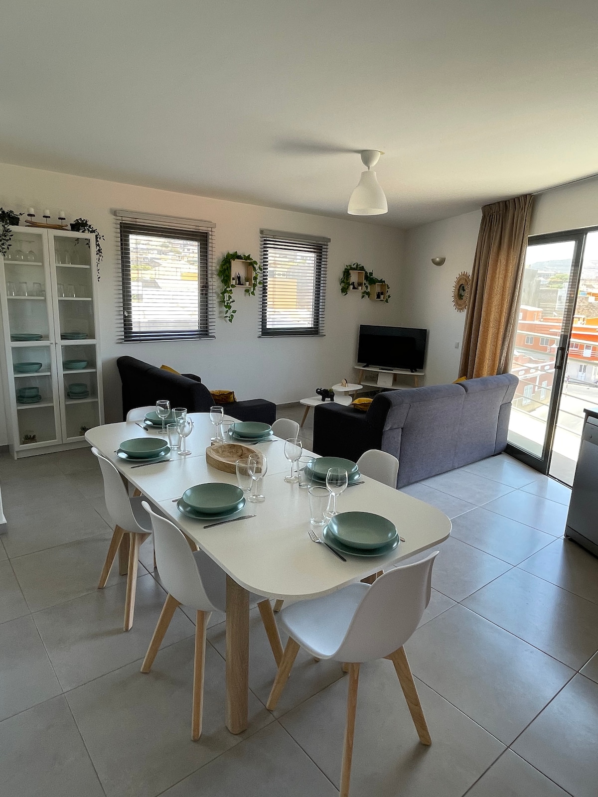 A bright dining area is featured, with a spacious white table set for six, complemented by light blue plates and glassware. Comfortable seating arrangements include a gray sofa and armchair. Large windows provide ample natural light, and a flat-screen TV is visible in the background.