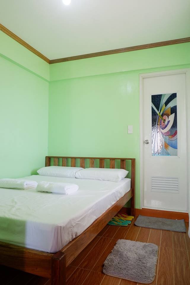 A simple bedroom is presented with light green walls and a wooden bed. The bed is covered with fresh linens and accompanied by rolled towels. A door with a stained glass design leads to the bathroom, creating an inviting space for relaxation.