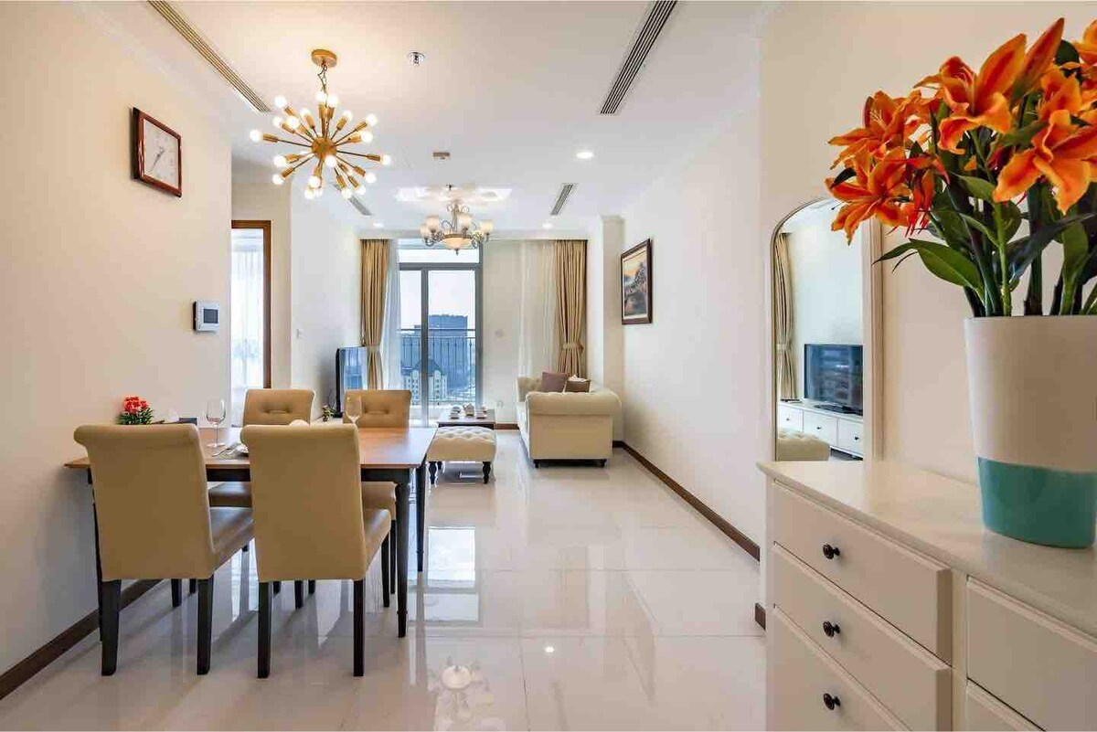 The living area is furnished with a dining table surrounded by four chairs, and a comfortable sofa facing a wall-mounted smart TV. A large window allows natural light to brighten the space, while decorative flowers add a touch of color on a nearby console.