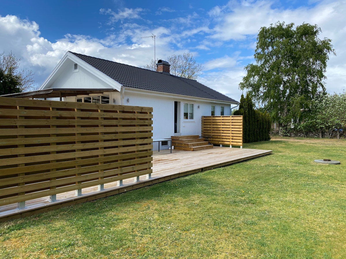 A single-story house is displayed, featuring a spacious wooden deck along the front. Surrounding greenery enhances the outdoor space, while wooden panels provide privacy. The exterior showcases a white facade with a black roof, under a partly cloudy sky.