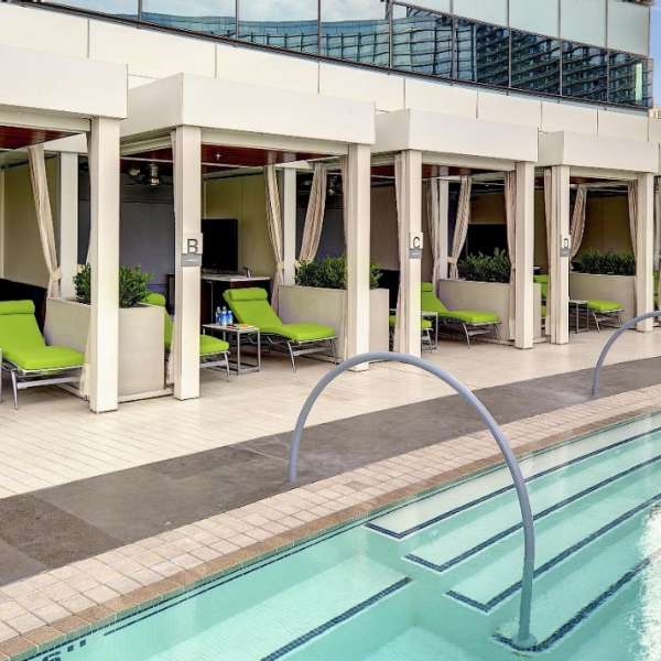 A serene rooftop pool area features a clean design, with several green lounge chairs positioned under shaded cabanas. The pool's gentle curves reflect the surroundings, while modern glass structures rise in the background.