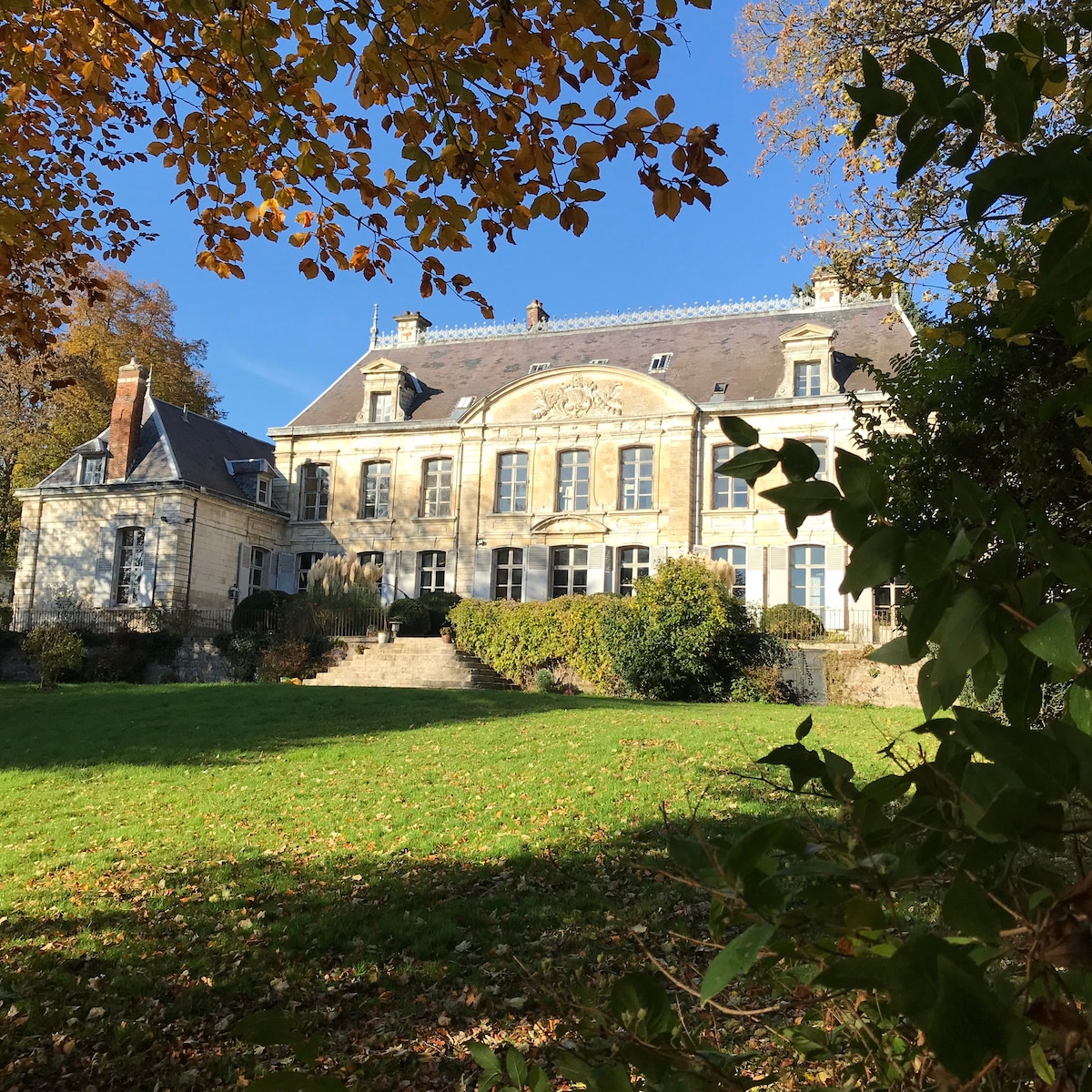 The exterior of the Chateau de Contay is framed by leafy trees, showcasing its elegant façade with tall windows and intricate architectural details. A lush green lawn leads to a set of stone steps that ascend to the entrance, highlighting the grandeur of the chateau.