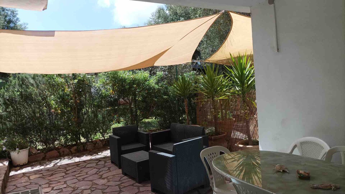 An outdoor area features shaded seating arrangements under a canvas awning. Potted plants introduce greenery, complementing the stone patio. A round dining table is positioned nearby, creating a relaxed atmosphere for outdoor dining or gatherings.