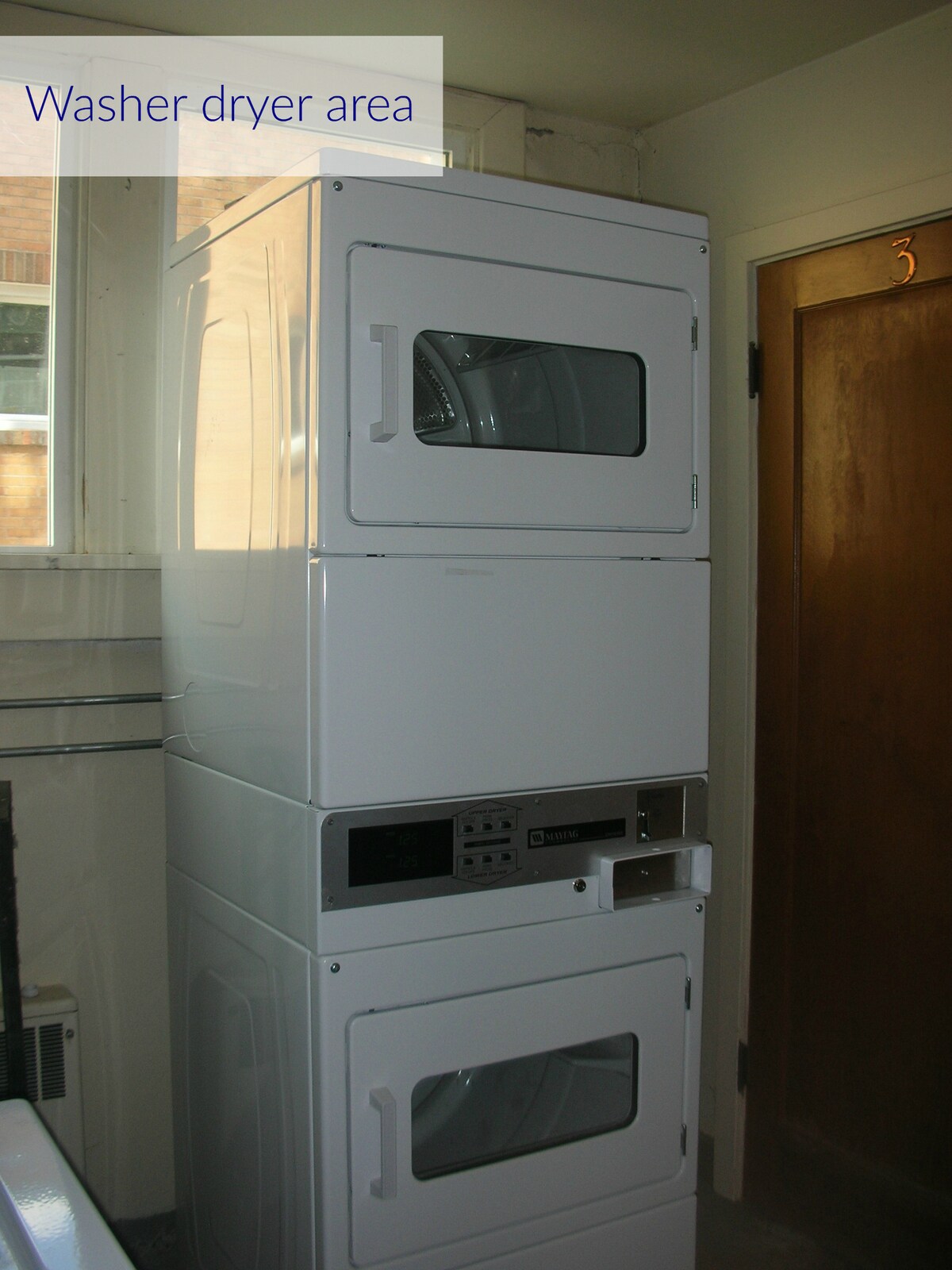 A stacked washer and dryer occupies a small laundry area, featuring a white exterior and front-loading doors. A window allows natural light into the room, and a wooden door with a number is visible nearby.