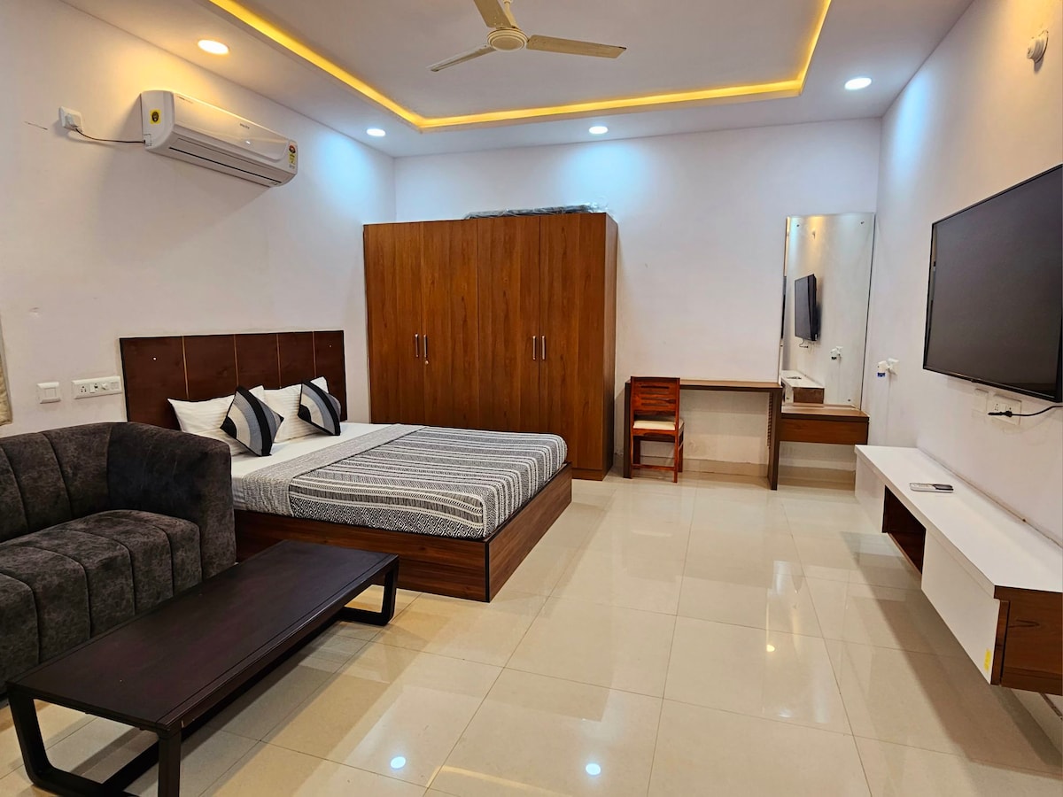 A spacious bedroom is presented, featuring a comfortable bed with a striped blanket. A plush seating area is included alongside a wooden wardrobe, desk, and mirror. A large television is mounted on the wall. Soft lighting and polished tiles enhance the modern aesthetic.