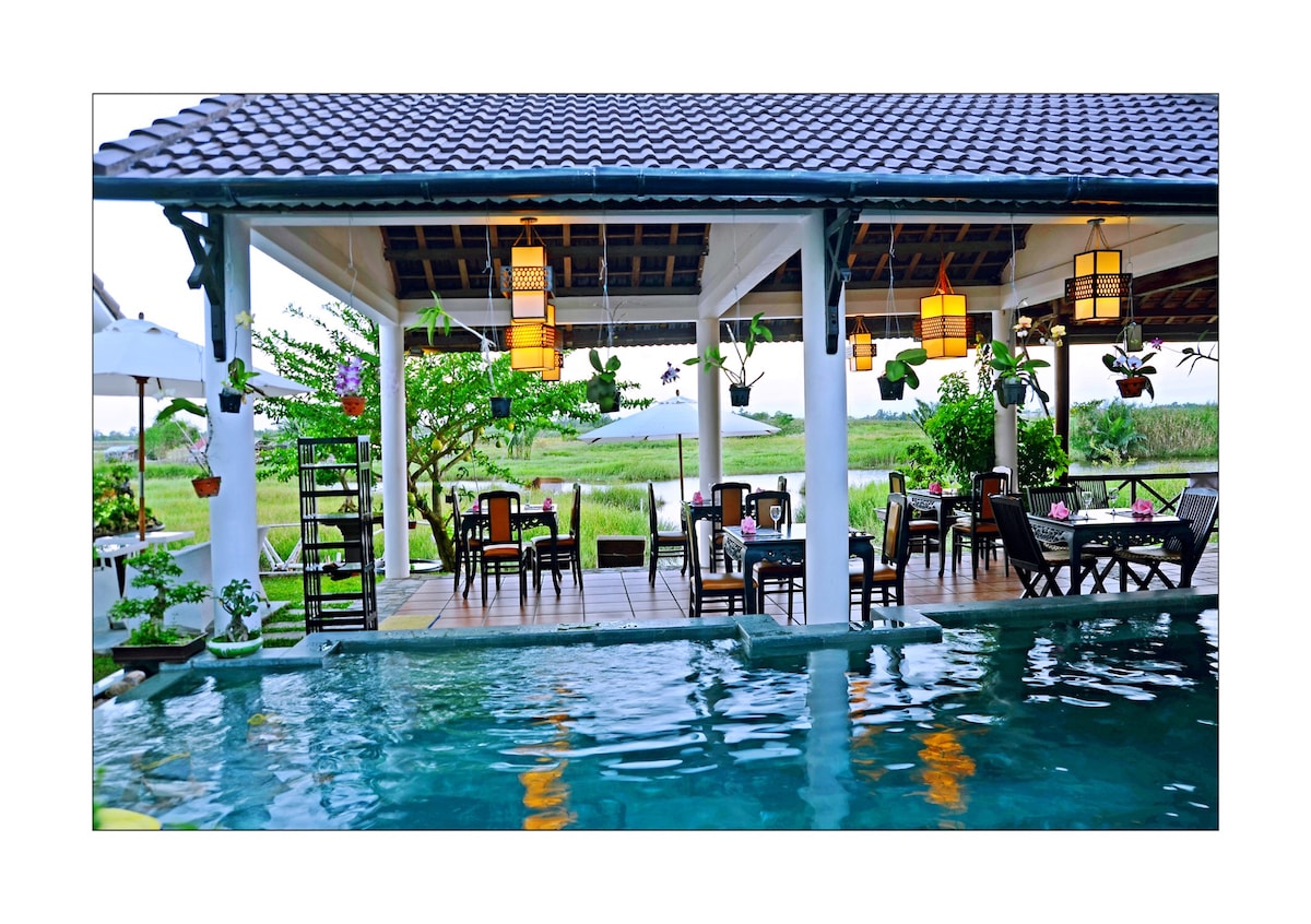 An open dining area features wooden tables and chairs surrounded by greenery, providing a relaxed setting. Lanterns hang from the ceiling, casting soft light over the space. A tranquil pool reflects the serene landscape, enhancing the peaceful ambiance.