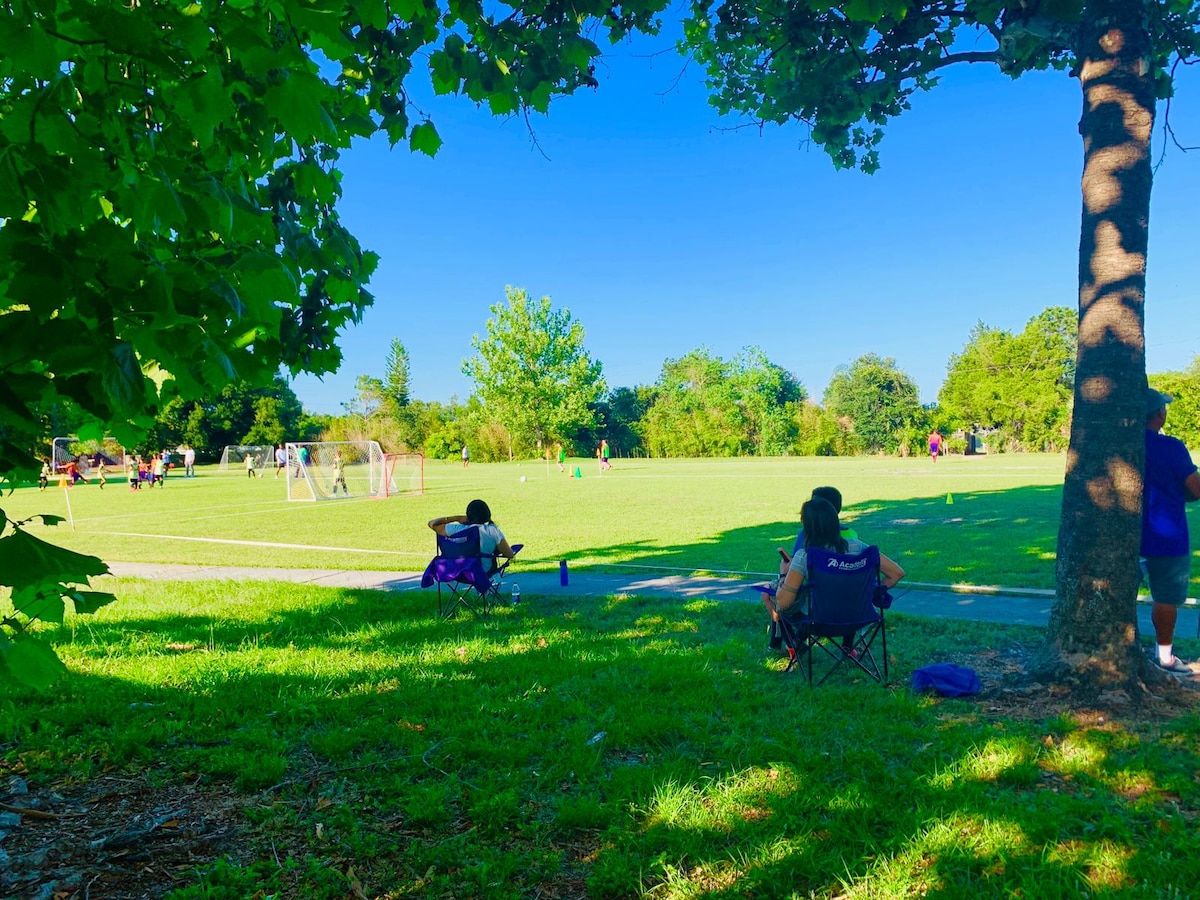 A spacious green park features a soccer field, with people engaged in various activities. In the foreground, two individuals relax in foldable chairs under a tree, enjoying the sunny day. Lush greenery surrounds the area, creating a peaceful outdoor setting.