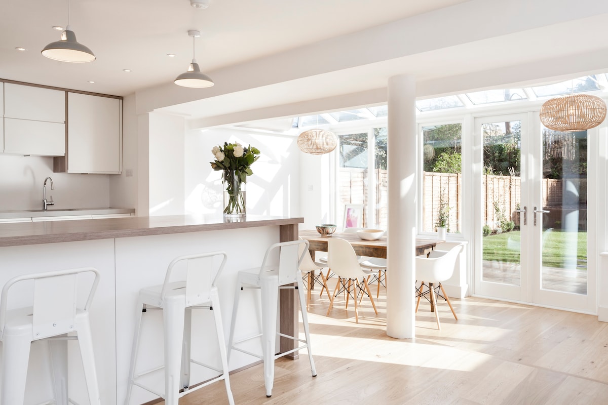 An open-plan kitchen and dining area features a modern design with a spacious kitchen island and four bar stools. Large glass doors lead to a garden, while natural light streams in through the conservatory's windows, illuminating the rustic dining table set for six.