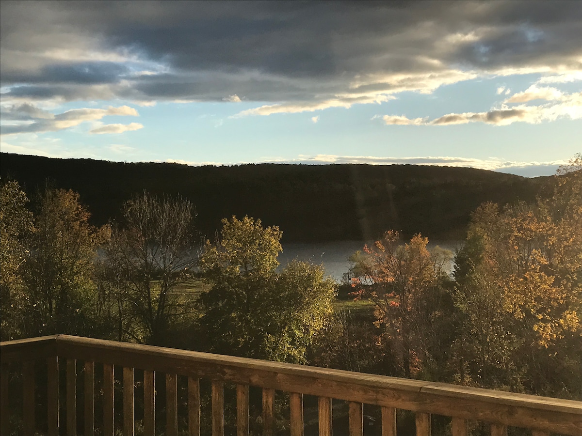 A scenic view from a wooden deck captures a tranquil lake surrounded by trees displaying vibrant fall foliage. Soft rays of sunlight break through the clouds, illuminating the landscape and highlighting the natural beauty of the area.