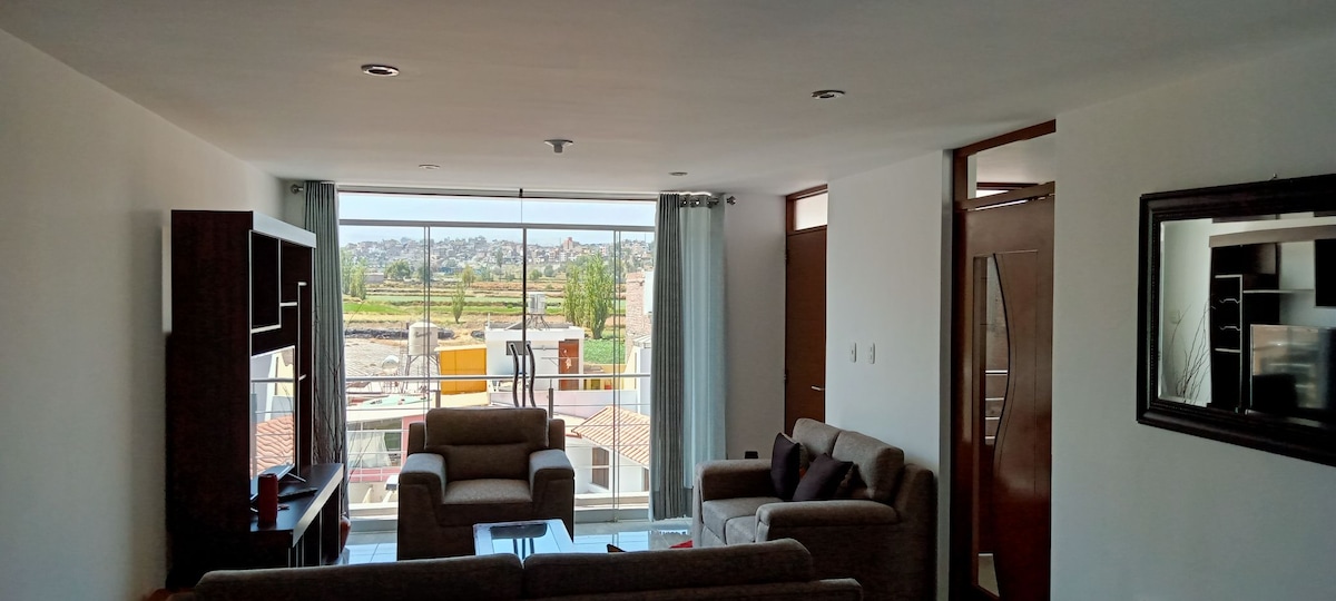 A bright living area is depicted with a large window showcasing a view of the surrounding landscape. Comfortable seating is provided by multiple sofas arranged thoughtfully, complemented by a coffee table in the center. Natural light fills the space, enhancing the open atmosphere.