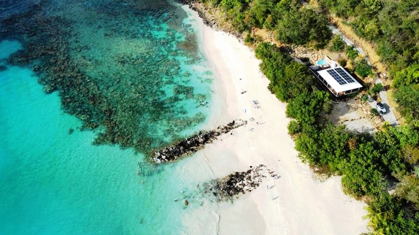 An aerial view presents a serene coastline meeting vibrant turquoise waters. Soft, powdered sand stretches along the beach, bordered by lush greenery. The villa is positioned nearby, surrounded by natural beauty, with rocky formations visible in the shallow water, enhancing the tranquil atmosphere.