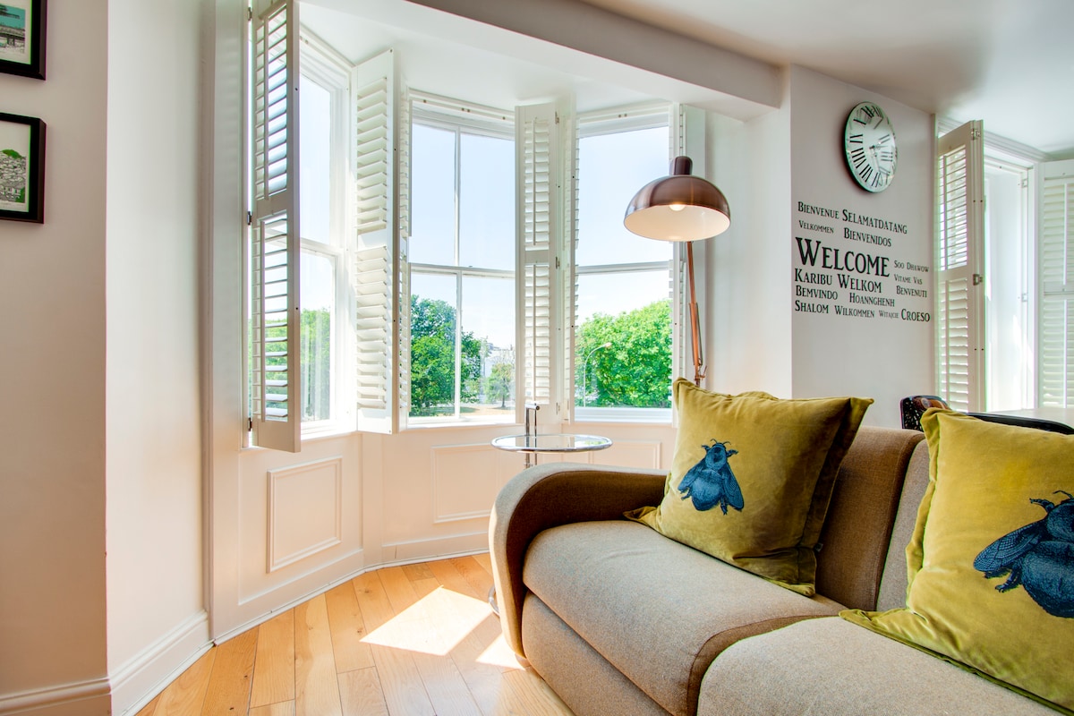 The open-plan lounge features a bay window with shuttered panels, allowing ample natural light. A comfortable sofa is complemented by decorative cushions. A small side table is positioned near the window, providing a cozy spot to relax and enjoy the view of the surrounding greenery.
