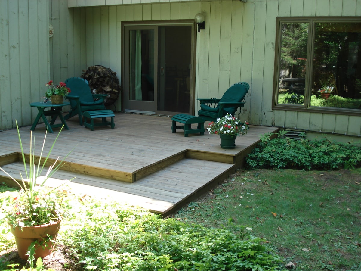 An outdoor deck is bordered by landscaped greenery and features a set of two wooden chairs alongside a small table. Potted flowers add a touch of color, while a stack of firewood is positioned nearby. Large windows provide views into the living area.