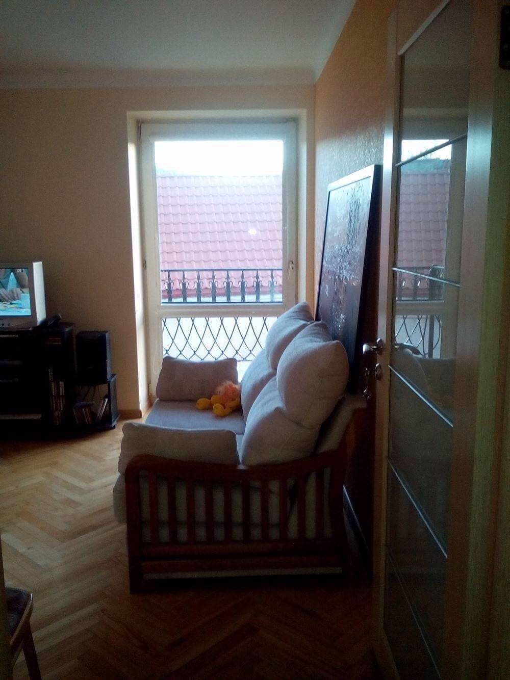 A cozy seating area features a light-colored couch positioned near a window, providing a view of the outside. A colorful stuffed toy is visible on the couch. Light-colored walls and parquet flooring create a warm atmosphere.