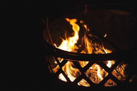 A fire pit is shown with flames flickering against the darkness of the night. The warm glow of the fire highlights the textured wood and metal structure surrounding it, creating a cozy ambience for gatherings.