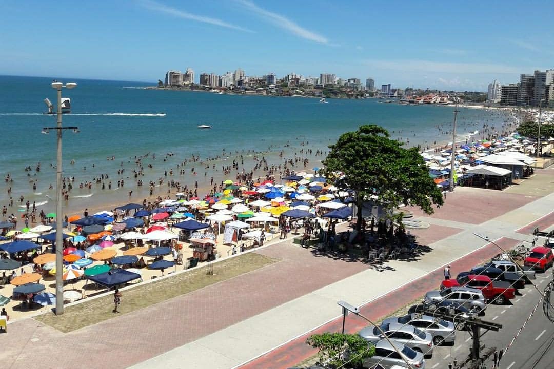 A vibrant beach scene is depicted, featuring numerous colorful umbrellas and beachgoers enjoying the sunny shore. The expansive ocean stretches into the distance, with buildings lining the coastline. A boardwalk runs parallel to the beach, accompanied by parked cars and palm trees.