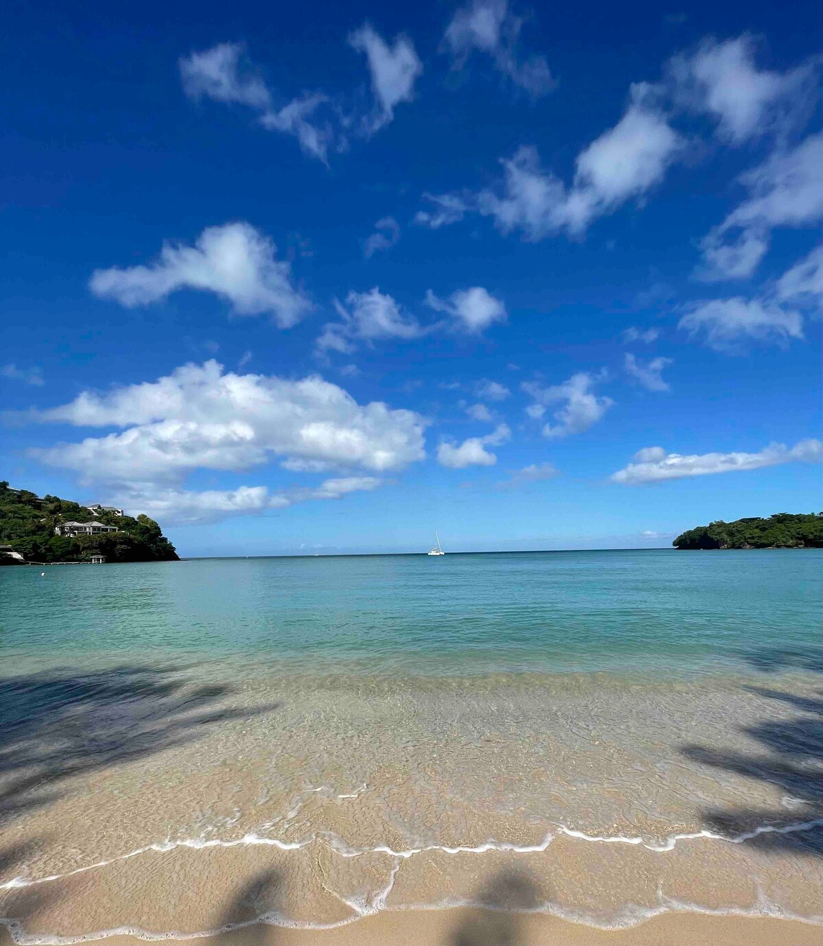 The image captures a tranquil beach scene, with clear blue waters gently lapping against a sandy shore. A sailboat is visible in the distance beneath a vast sky adorned with fluffy white clouds, while lush green hills frame the beach on either side.