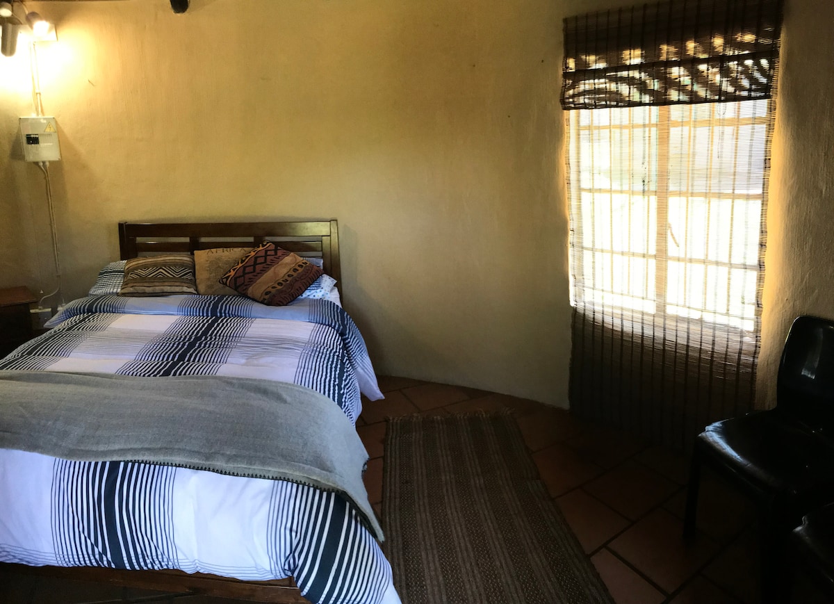 A cozy sleeping area features a wooden bed with a patterned duvet cover and decorative pillows. Natural light filters through a window with bamboo shades, illuminating the room's warm tones. A simple chair and a woven rug add to the comfortable ambiance.