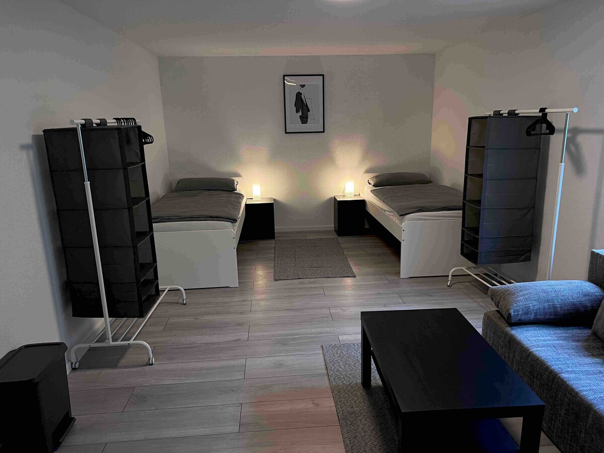 The room includes two spacious beds positioned parallel to each other, with nightstands on either side. A cozy seating area features a gray sofa, and a low black coffee table sits nearby. Soft illumination from lamps enhances the minimalist decor.
