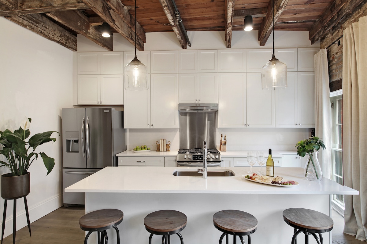 An open-concept kitchen with modern finishes features a large island with seating for several guests. Stainless steel appliances complement white cabinetry, while wooden beams add warmth to the ceiling. Natural light streams in through a window, enhancing the inviting space.
