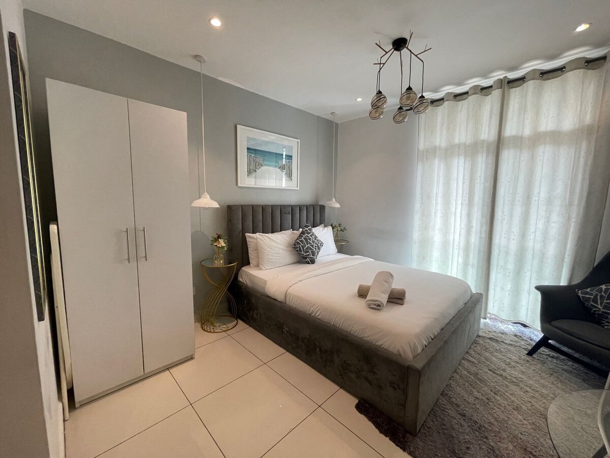 A cozy room features a spacious bed adorned with neatly arranged towels. A stylish wardrobe and a decorative side table complement the design. Natural light filters through sheer curtains, creating a soft ambiance in the inviting space. A comfortable chair adds to the room's functionality.