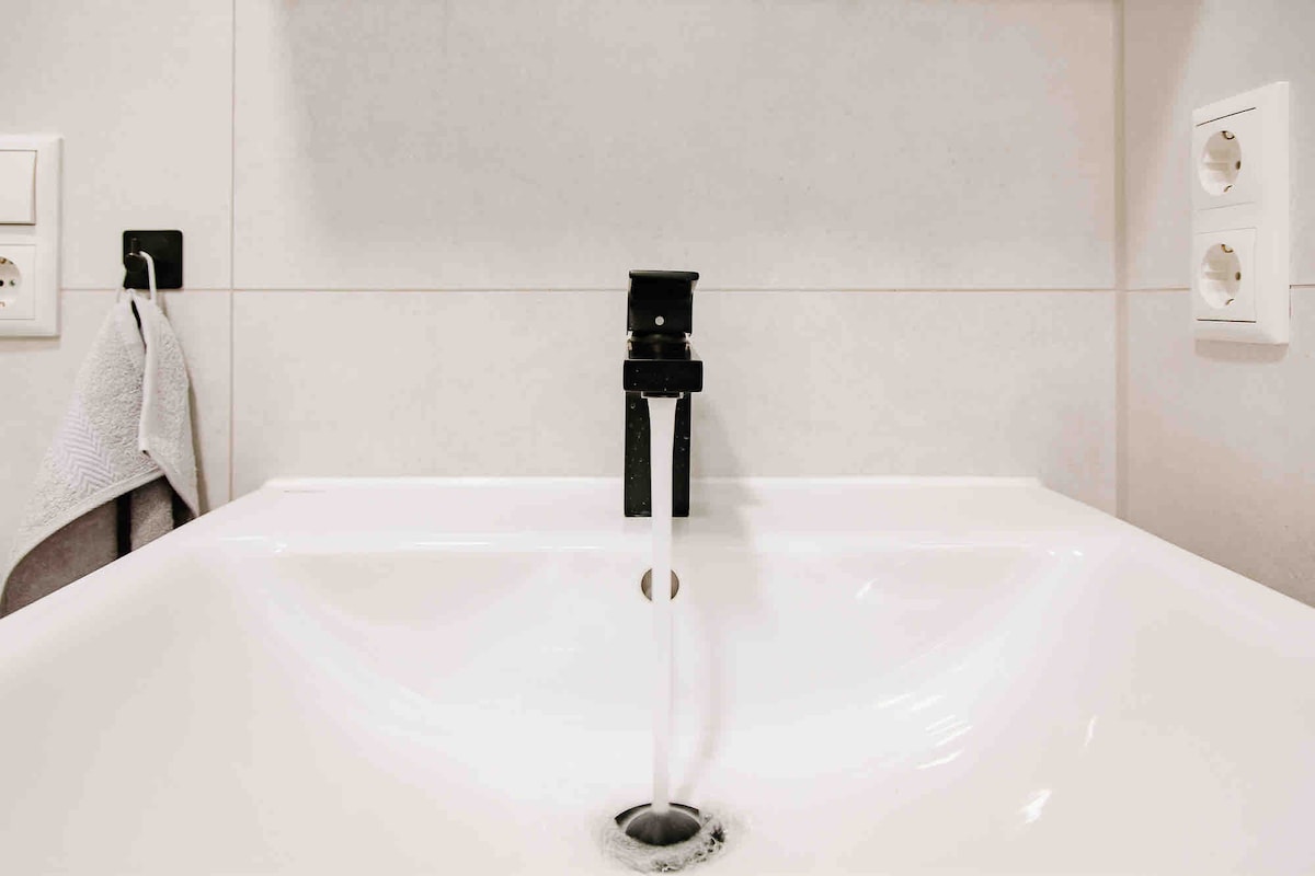A modern sink is displayed, featuring a sleek black faucet from which water is flowing into the basin. Light-colored tiles surround the sink, and a towel is hanging nearby, enhancing the functional yet minimalist design.
