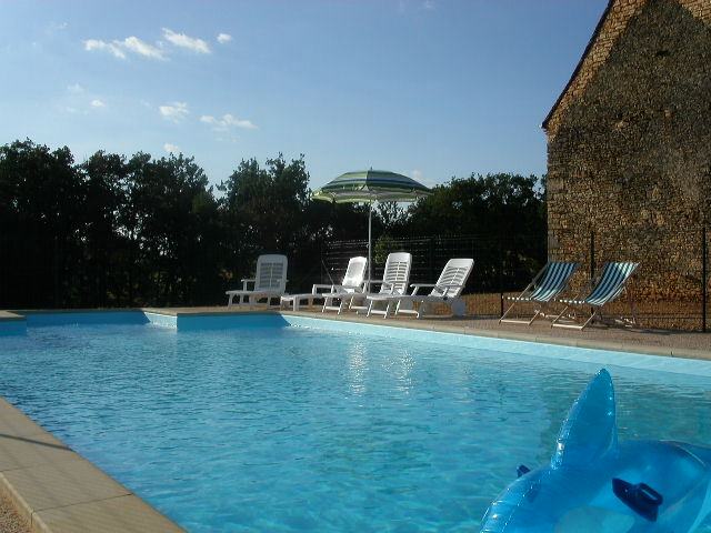 A private swimming pool features clear blue water surrounded by lounge chairs and a sun umbrella. The pool area is bordered by a stone wall and greenery, offering a serene outdoor space for relaxation.
