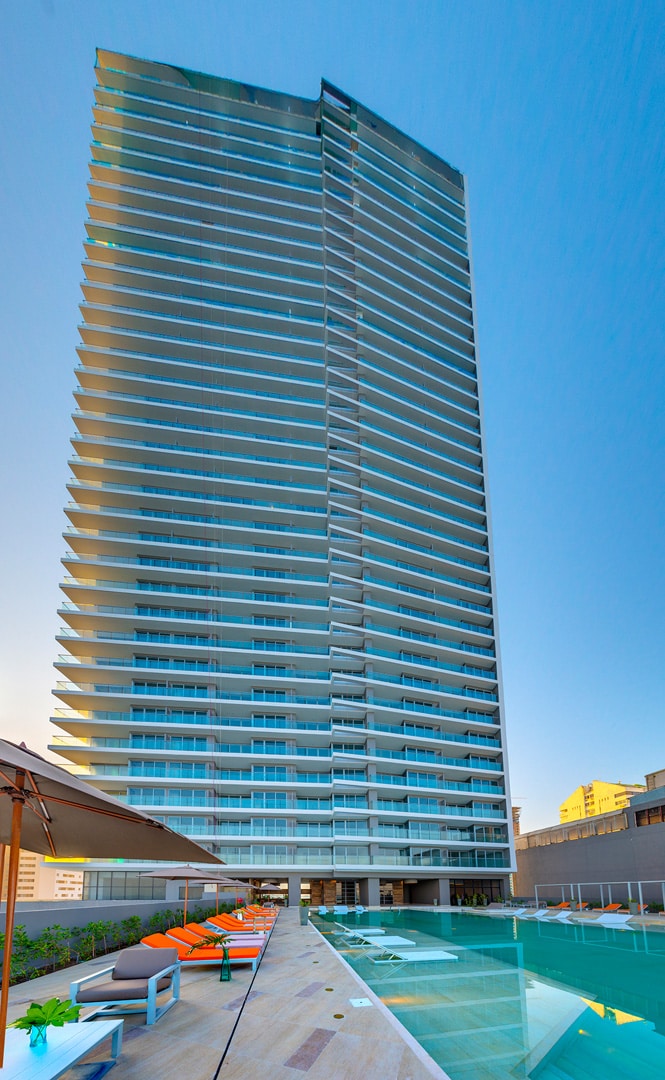 A modern high-rise building features a sleek design, showcasing many glass balconies and large windows. The structure stands above a landscaped area, with lounge chairs and umbrellas arranged along the poolside, inviting relaxation.