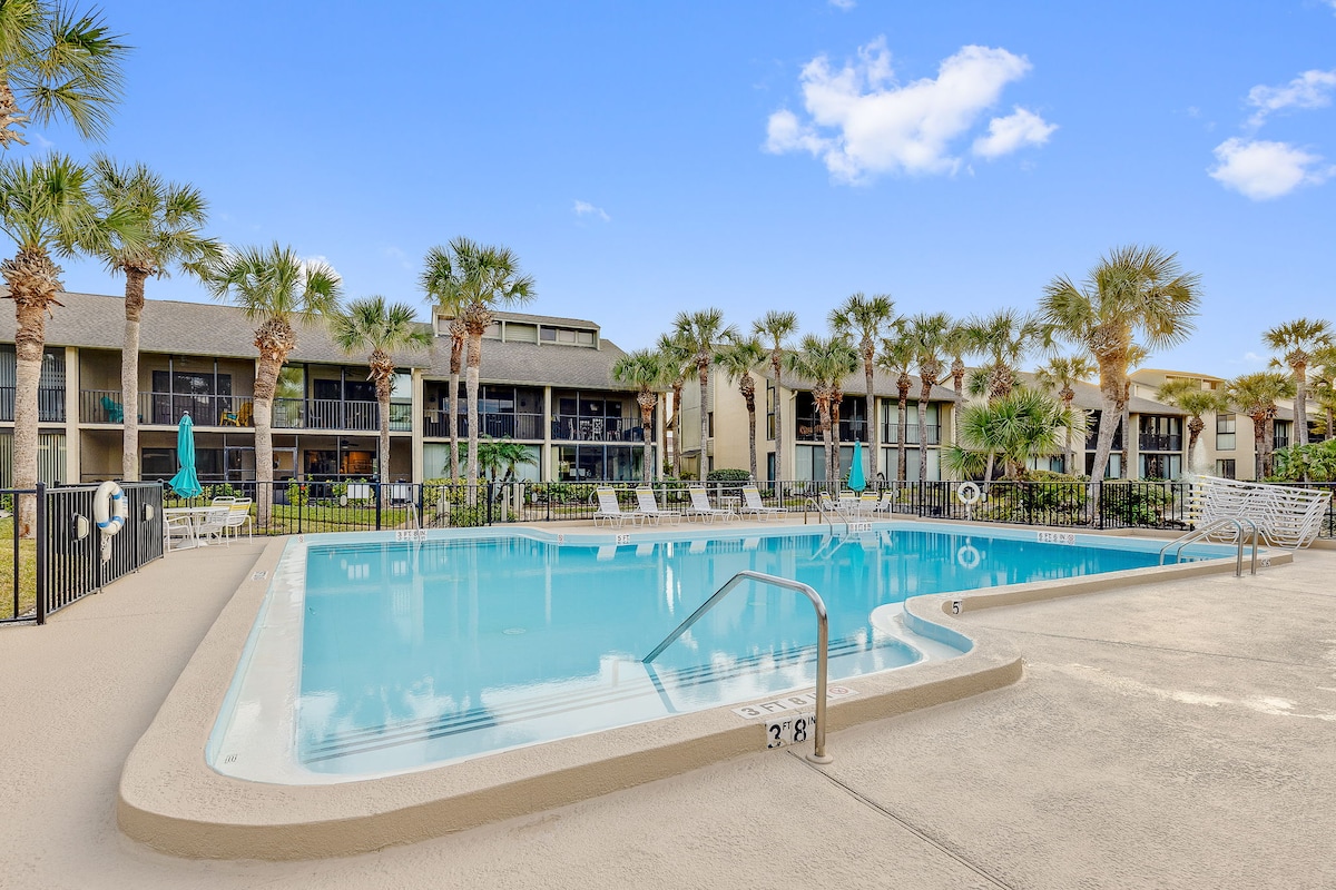 An outdoor pool area surrounded by palm trees provides a serene setting for relaxation. Lounge chairs are positioned around the pool, with a gated property visible in the background. The clear blue sky enhances the inviting atmosphere of the resort community.