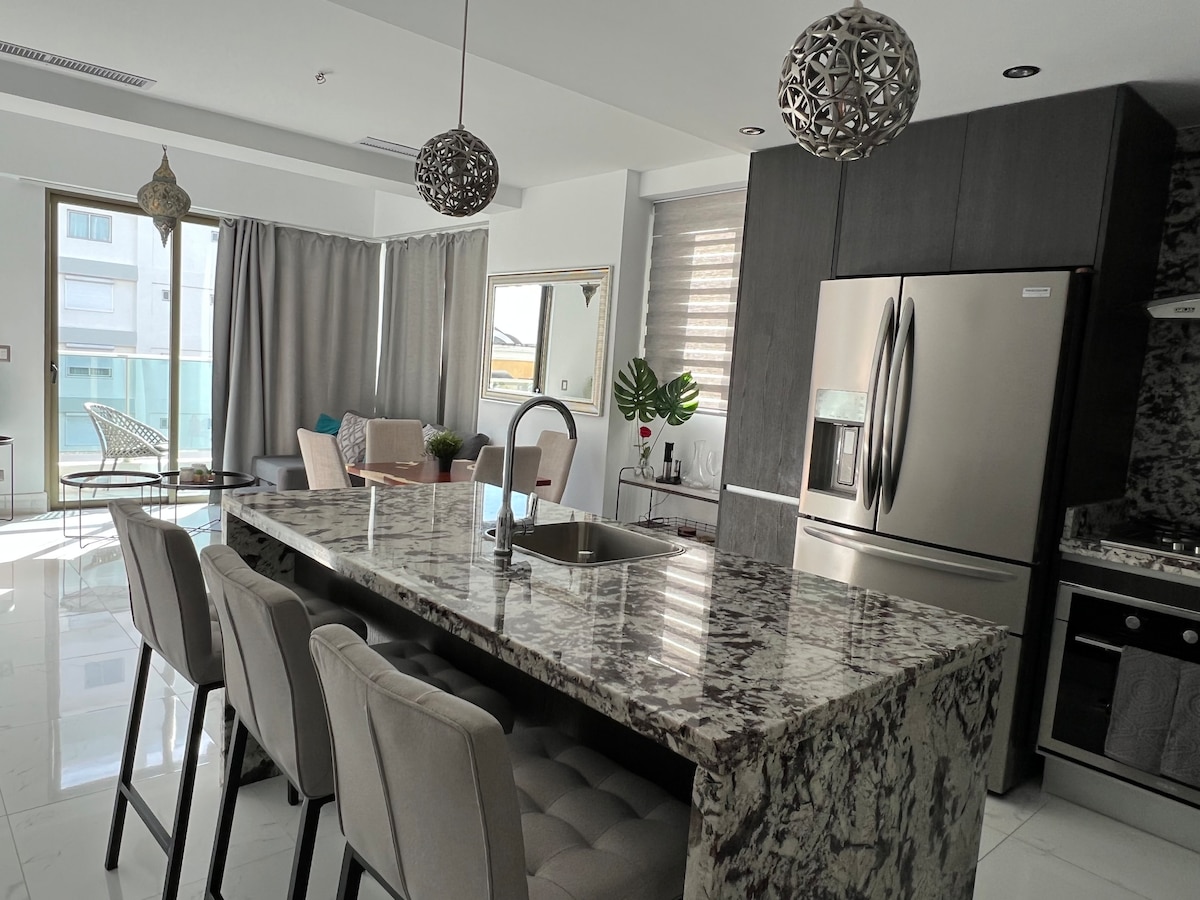 A modern kitchen and living area are highlighted by an expansive marble island with seating. Large windows allow natural light to flood the space, complemented by sleek cabinetry and stylish pendant lights. Soft seating is arranged nearby, enhancing the open layout.