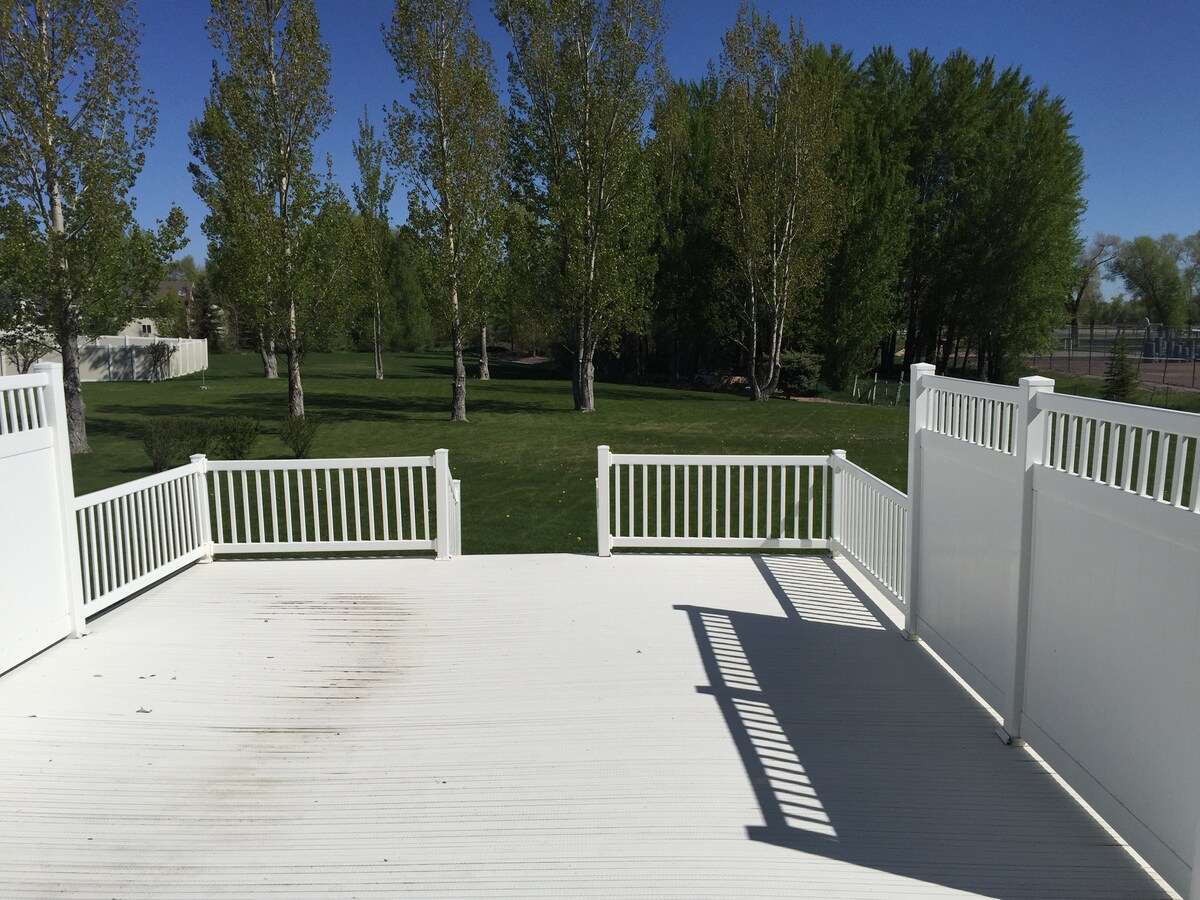 A spacious outdoor deck is presented, featuring white railing and flooring. The surrounding greenery is visible, including tall trees and an expansive lawn. Natural light creates a bright atmosphere, while shadows from the railing add visual interest to the space.
