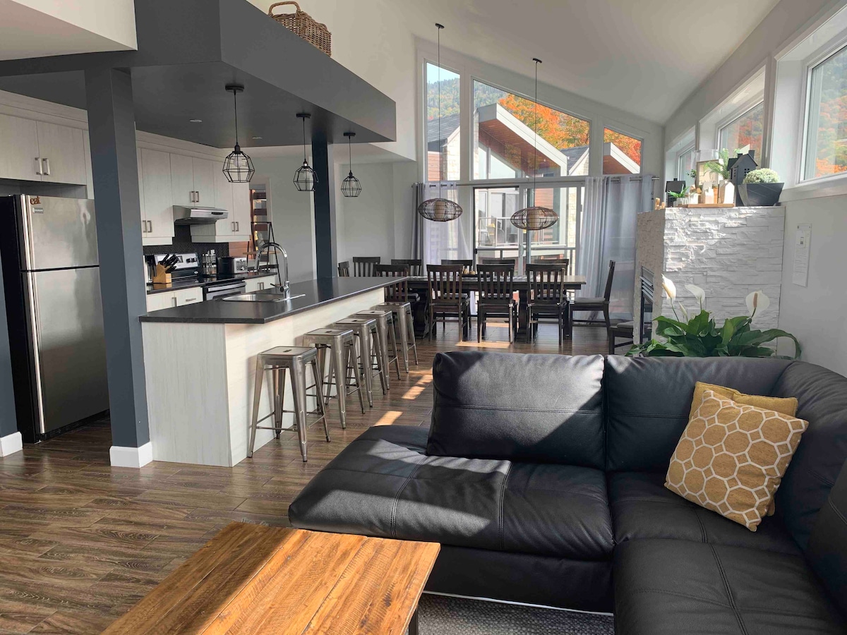 An open living space features a modern kitchen with stainless steel appliances and a sleek island bar surrounded by metal stools. Sunlight pours through large windows, illuminating the dining area equipped with a long wooden table and matching chairs. A cozy sofa with a decorative pillow invites relaxation.
