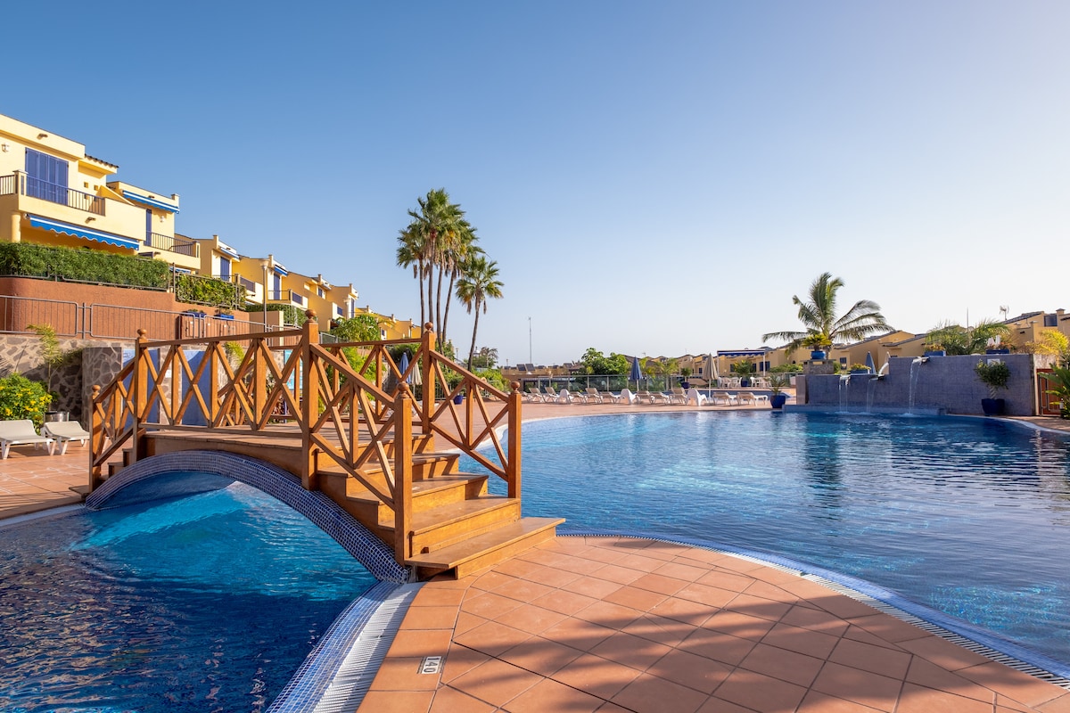 A tranquil pool area features a wooden bridge arching over the water. Lush palm trees provide greenery alongside the pool, which is surrounded by tiled paths and sun loungers. The clear blue sky reflects on the calm water, enhancing the inviting atmosphere of the outdoor space.