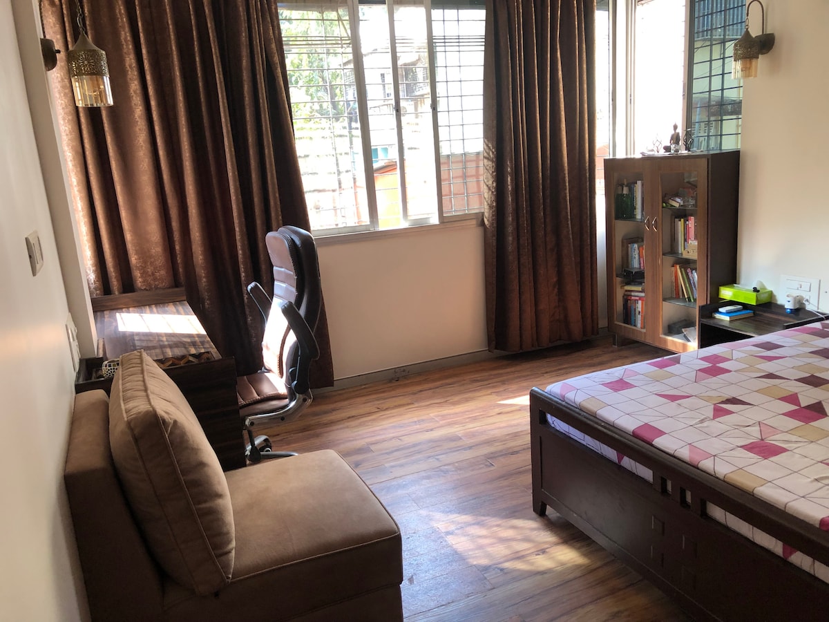 A well-lit bedroom features large windows dressed in brown curtains, allowing natural light to brighten the space. A spacious bed with a patterned bedspread is accompanied by a cozy sofa. A wooden desk with an ergonomic chair and a small bookshelf filled with books enhance the functional layout.
